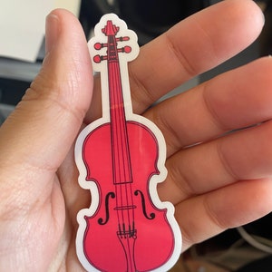 Puede incluir: Una pegatina de viol&iacute;n roja con contornos y detalles negros. La pegatina est&aacute; sobre un fondo blanco.