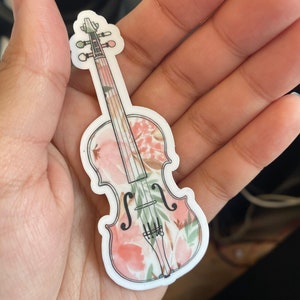 Puede incluir: Una pegatina de vinilo blanco de un viol&iacute;n con un patr&oacute;n floral en tonos de rosa y verde.