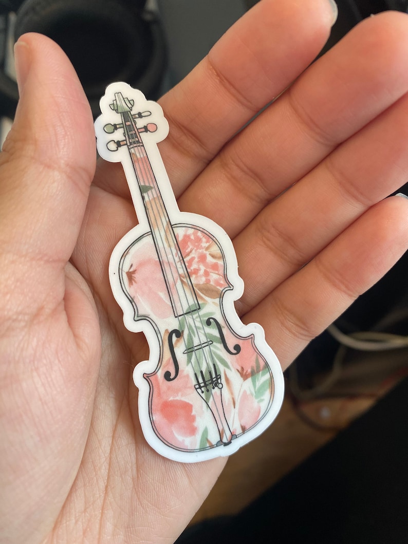Puede incluir: Una pegatina de vinilo blanco de un viol&iacute;n con un patr&oacute;n floral en tonos de rosa y verde.
