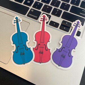 Puede incluir: Tres pegatinas de vinilo de violines en diferentes colores: azul, rojo y morado. Los violines son todos del mismo estilo y tienen un dise&ntilde;o sencillo.