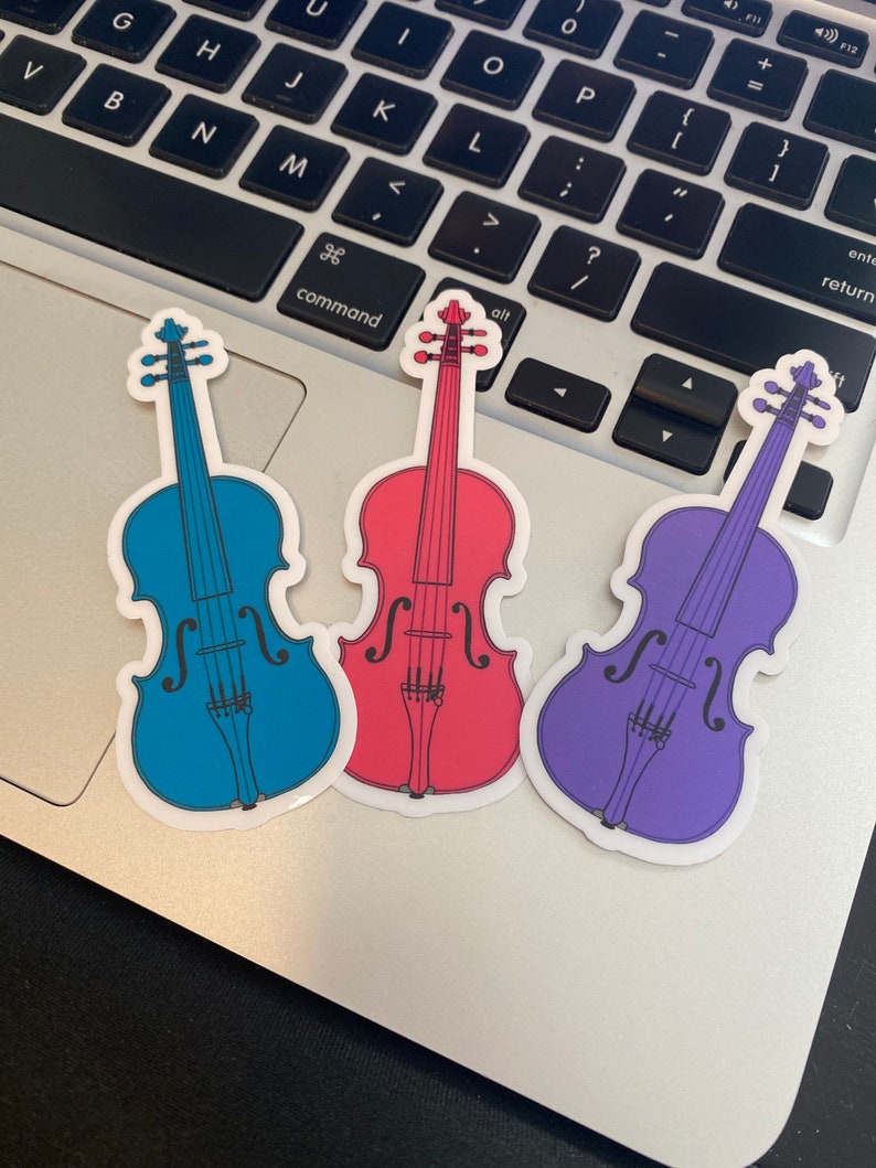 Puede incluir: Tres pegatinas de vinilo de violines en diferentes colores: azul, rojo y morado. Los violines son todos del mismo estilo y tienen un dise&ntilde;o sencillo.