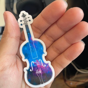 Puede incluir: Una pegatina de vinilo de un viol&iacute;n con un dise&ntilde;o de galaxia. El viol&iacute;n est&aacute; contorneado en blanco y el dise&ntilde;o de la galaxia est&aacute; en tonos de azul, morado y rosa.