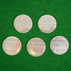 May include: Five round, white, ceramic coasters with a printed design of vintage sheet music. The sheet music is printed in black and gray ink.