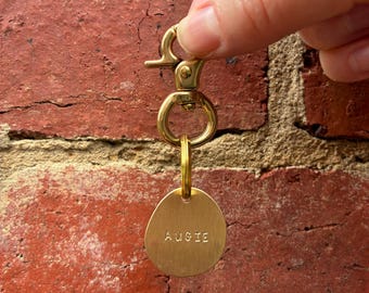 Peut inclure: Un porte-clés en métal doré composé d'un fermoir mousqueton relié à un anneau circulaire, lui-même attaché à un petit pendentif en laiton de forme irrégulière. Le pendentif porte l'inscription AUGIE gravée en lettres capitales sur sa face.