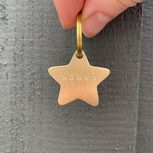 May include: A gold-coloured star-shaped dog tag with the word "HOUND" engraved on it. The tag is attached to a gold-coloured ring. The background is a grey wooden surface.
