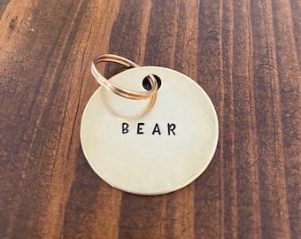 May include: A round, gold-colored metal tag with a key ring attached. The tag has the word "BEAR" engraved on it.