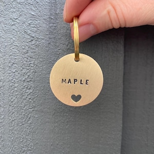 Puede incluir: Una etiqueta redonda de metal dorado con el nombre "MAPLE" estampado en letras negras. Un recorte en forma de corazón está debajo del nombre. La etiqueta está unida a un anillo dorado.