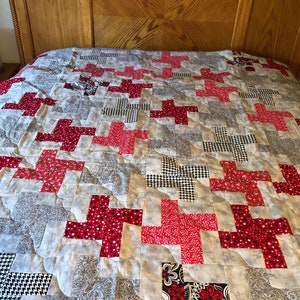 Op de afbeelding: Een rood en grijs quilt met een kruispatroon. De quilt is gemaakt van verschillende stoffen met verschillende patronen, waaronder stippen, ruitjes en bloemenpatronen.
