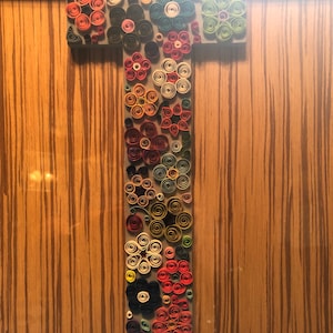 Può includere: Un'opera d'arte in quilling di carta colorata a forma della lettera T. La lettera è composta da molti piccoli fiori di carta arrotolati in vari colori, tra cui rosso, arancione, giallo, verde, blu e viola.
