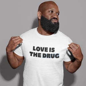 May include: White t-shirt featuring the text "LOVE IS THE DRUG" in bold, black lettering. The word "DRUG" is underlined in light blue. The shirt is worn by a person with a shaved head and a dark, full beard.