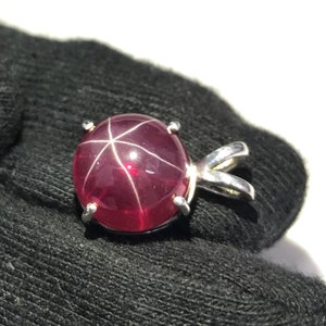 Könnte beinhalten: Ein roter Sternrubin-Anhänger mit einer silbernen Fassung. Der Rubin ist im Cabochon-Schliff geschliffen und hat ein sternförmiges Muster.