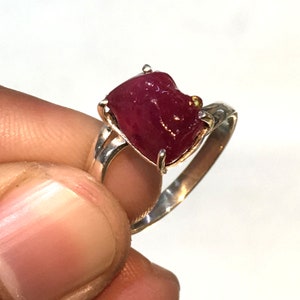 Puede incluir: Un anillo de plata con una gran piedra preciosa de rubí rojo en bruto. El rubí está engastado en garras y tiene una pequeña piedra preciosa amarilla en el lateral.