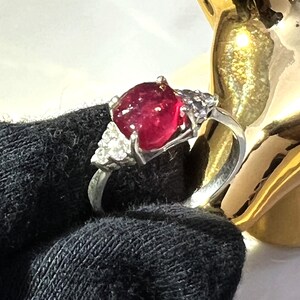 Puede incluir: Un anillo de plata con una piedra preciosa roja grande de forma ovalada rodeada de piedras preciosas más pequeñas y transparentes.