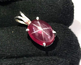 Puede incluir: Un colgante de rubí estrella roja con un engaste de plata. El rubí tiene forma ovalada y presenta un patrón en forma de estrella en el centro.