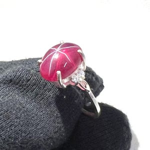 May include: A silver ring with a large, red star ruby gemstone and smaller white gemstones on the band.