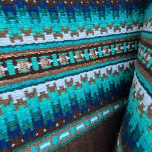 Pode incluir: Um close-up de um tecido tecido marrom e azul com um padr&atilde;o geom&eacute;trico. O tecido tem uma faixa branca que atravessa o centro e &eacute; feito de um material macio e texturizado.