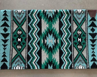 May include: A turquoise, green, black, and white saddle pad with a geometric pattern. The saddle pad has a brown leather patch on the right side.