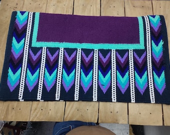 May include: A rectangular saddle pad with a dark purple center and a teal border. The pad features a repeating pattern of teal, purple, and blue chevrons separated by white vertical lines. The edges are dark blue.