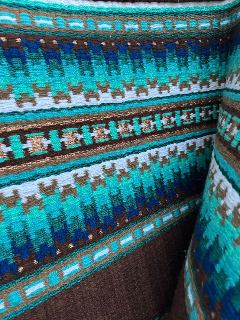 Pode incluir: Um close-up de um tecido tecido marrom e azul com um padr&atilde;o geom&eacute;trico. O tecido tem uma faixa branca que atravessa o centro e &eacute; feito de um material macio e texturizado.
