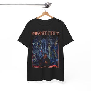 以下が含まれることがあります： 「NIGHT CITY」という赤いオレンジ色の文字が書かれた、黒いTシャツ。都市の風景が描かれており、ネオンライトが輝く高い建物の前に人が立っている様子が描かれている