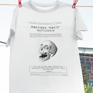 Puede incluir: Camiseta blanca con una ilustración en blanco y negro de una calavera con la boca abierta y un ojo visible. El texto "Mortimer "Morte" Rictusgrin" está impreso sobre la calavera. Debajo de la calavera hay una cita: "Eres tú y yo, berk - estamos en esto juntos. Hasta que esto termine. Me quedo como tu pierna." - Mor+E-