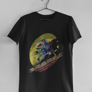 Puede incluir: Camiseta negra con un estampado gráfico de cuatro personajes de dibujos animados en una azotea frente a una luna amarilla. Los personajes llevan ropa azul, verde y roja.