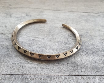 May include: A gold-toned cuff bracelet with a series of small, dark triangle patterns. The open-ended bracelet is slightly curved and rests on a weathered, gray wooden surface. The bracelet's design is simple yet elegant.