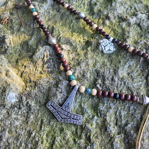Könnte beinhalten: Eine Holzperlenkette mit einem silbernen Mjolnir-Anhänger und einem silbernen Herz-Anhänger. Die Perlen sind braun, schwarz und grün.