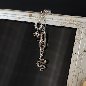 May include: A handheld gaming console with a white celestial-themed skin featuring gold sun and moon motifs. A silver-toned metal charm featuring a snake, a star, and a small pearl ring hangs from the top edge. A tan paw-shaped joystick cover is attached.