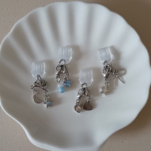 Può includere: Quattro ciondoli d'argento con cime in plastica trasparente. Ogni ciondolo presenta un design diverso: una luna crescente, una stella, un cuore e una piccola gemma blu. I ciondoli sono attaccati ad anelli di plastica trasparente.