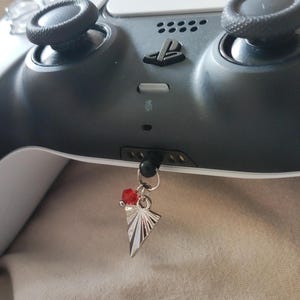 May include: A silver charm with a geometric design and a red bead hangs from a black PlayStation 5 controller. The controller is primarily gray and black.