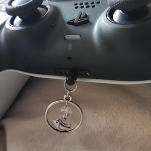 May include: A silver charm with a bird design and a clear crystal bead hangs from a black PlayStation controller. The charm is circular and has a small bird silhouette inside. The controller is dark gray.