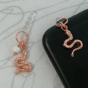 May include: Rose gold phone charm with a snake pendant and a white bead. The charm is attached to a clear plug. The snake pendant has a textured surface and is curled into an S-shape. The charm is on a black phone.