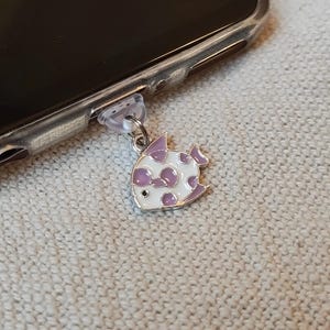 May include: A white and purple fish charm with a silver metal loop. The charm is attached to a clear plastic phone charger plug.