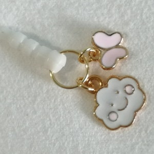 Op de afbeelding: Een witte oortelefoonaansluiting-bedel met een goudkleurige ring, met een roze en witte bloem en een witte wolk met een lachend gezicht. De bedel is ontworpen voor gebruik met een 3,5 mm audio-aansluiting.