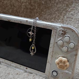 以下が含まれることがあります： 天体デザインの、ポータブルゲーム機用の透明保護ケース。星と涙のペンダントが付いたシルバーチェーンのチャームが上部から吊り下がっています。デバイスには白いボタンと肉球のジョイスティックカバーが付いています。