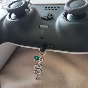 May include: A silver snake charm with clear crystals and a green bead hangs from a black PlayStation 5 controller. The controller is gray and black, with black joysticks and the PlayStation logo.