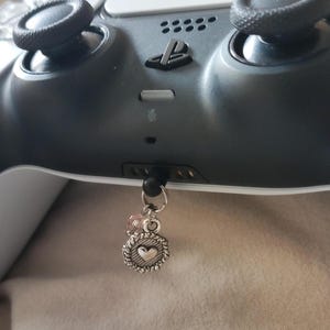 May include: A gray and white video game controller with a small silver charm attached. The charm features a heart design within a decorative border, accented with pink beads. The controller has black joysticks.