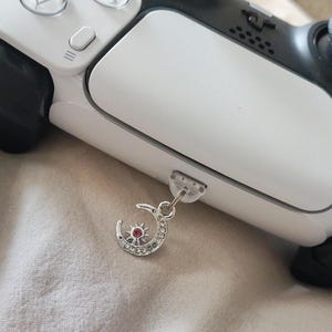 May include: A silver charm featuring a crescent moon and sun design with a red center stone, attached to a white and black game controller. The charm is detailed with small clear stones.