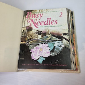 Könnte beinhalten: Ein Vintage-Magazin mit dem Titel Busy Needles 2, das auf dem Cover ein hellviolettes Stoffblumen-Accessoire mit grünen Blättern zeigt, das neben verschiedenen Kosmetikartikeln auf einer dunklen Oberfläche liegt. Der Text lautet What to make How to make it.