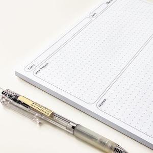 May include: A white notepad featuring a dot grid and sections for topic, key terms, date, and notes. A clear pen with a yellow label lies on the notepad. The notepad is designed for organisation and note-taking.