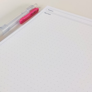 May include: A blank white page with a dotted grid pattern. The page has two lines at the top with the words "TOPIC" and "NOTES" written on them. A clear pen with a pink clip is lying on the page.