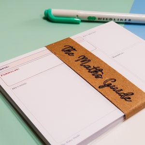 May include: A brown paper band with the text "The Maths Guide" wrapped around a white notebook with sections for topic, formulae, method, key notes, and example. The notebook is on a light green surface.
