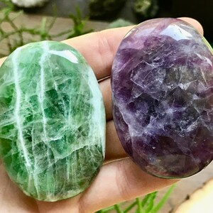 Könnte beinhalten: Zwei polierte, oval geformte Steine. Einer ist leuchtend grün mit weißen Adern, der andere tiefviolett mit helleren violetten und grauen Mustern. Die Steine werden in einer Hand gehalten, wodurch ihre glatten, glänzenden Oberflächen zur Geltung kommen.