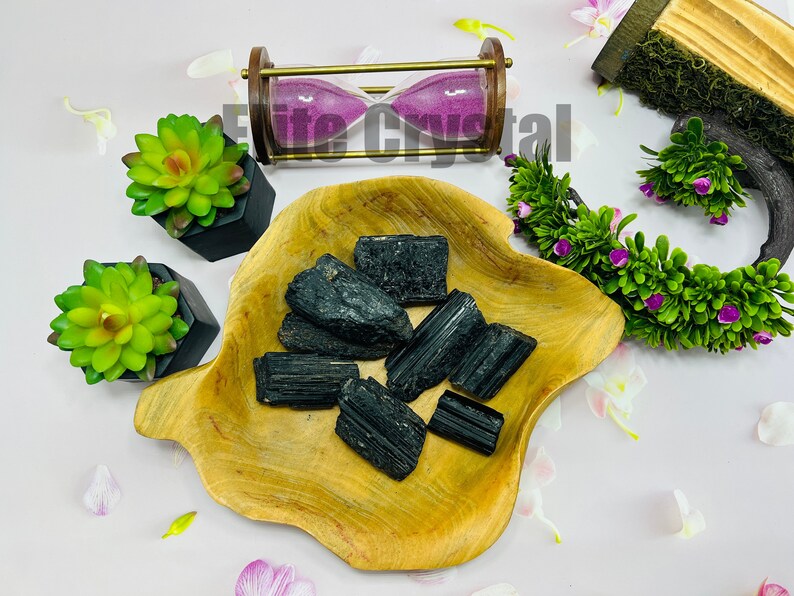 May include: A collection of raw, black tourmaline crystals displayed in a wooden bowl. The crystals have a rough, textured surface. The arrangement is complemented by decorative elements, including a miniature hourglass and artificial succulents.