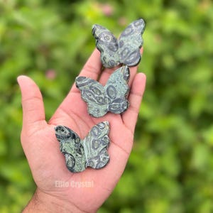 May include: Three green and gray stone butterfly figurines are held in a hand. The butterfly carvings have detailed wing patterns. The background is a blurred green, suggesting foliage. The text "Elite Crystal" is visible.