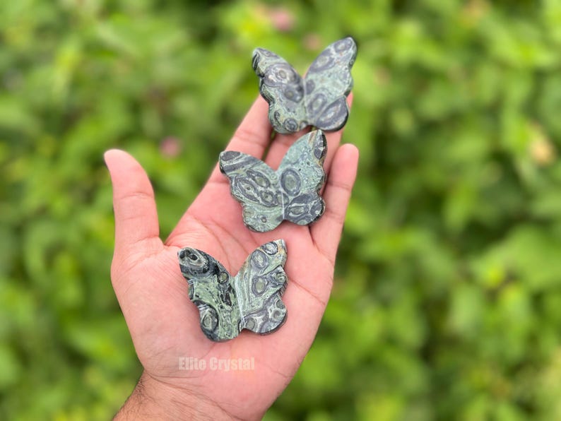 May include: Three green and gray stone butterfly figurines are held in a hand. The butterfly carvings have detailed wing patterns. The background is a blurred green, suggesting foliage. The text "Elite Crystal" is visible.