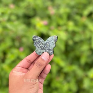 May include: A butterfly-shaped stone carving in shades of green and black is held up against a blurred green background. The stone has a detailed pattern and is held in a hand. The product is from Elite Crystal.