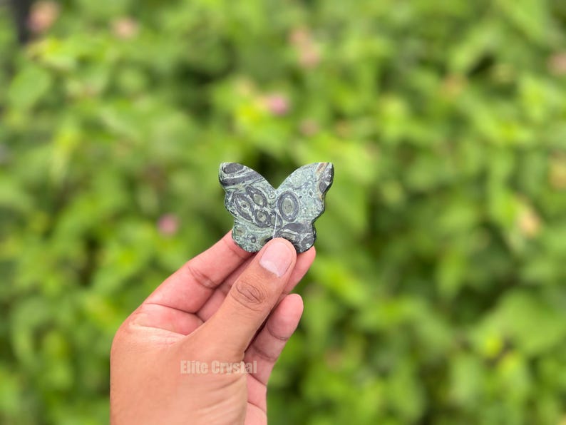 May include: A butterfly-shaped stone carving in shades of green and black is held up against a blurred green background. The stone has a detailed pattern and is held in a hand. The product is from Elite Crystal.
