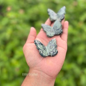 May include: Three green and black butterfly figurines are held in an open hand. The butterfly sculptures have detailed wing patterns. The background is a blurred green. The text "Elite Crystal" is visible at the bottom.